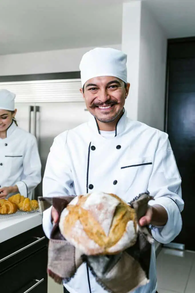 Smiling head chef presenting a freshly baked artisan loaf in a professional kitchen, representing culinary career success.