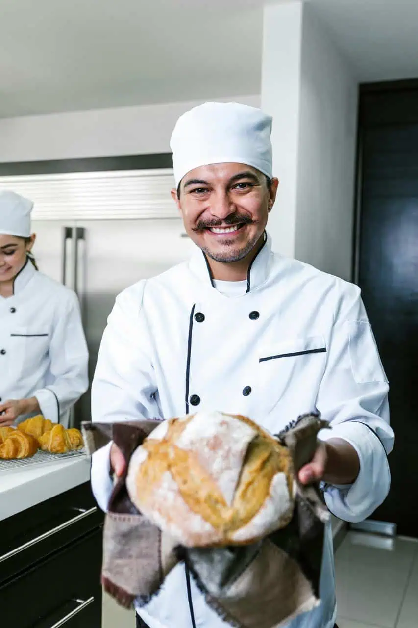 Smiling head chef presenting a freshly baked artisan loaf in a professional kitchen, representing culinary career success.