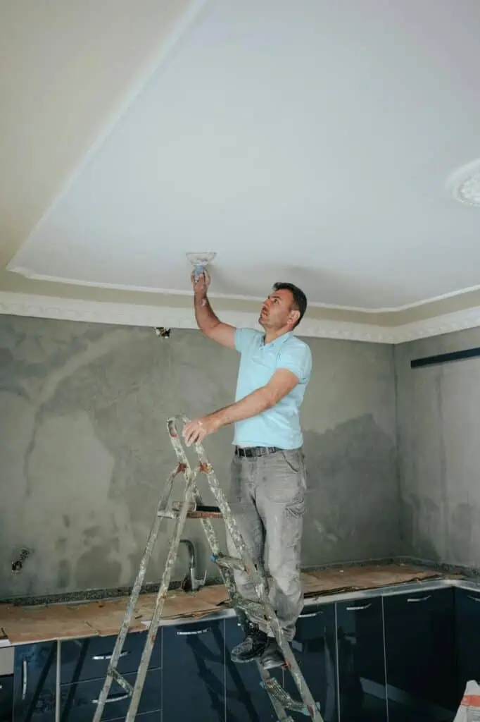 A hotel maintenance technician standing on a ladder, repairing a ceiling light fixture in a modern guest room.