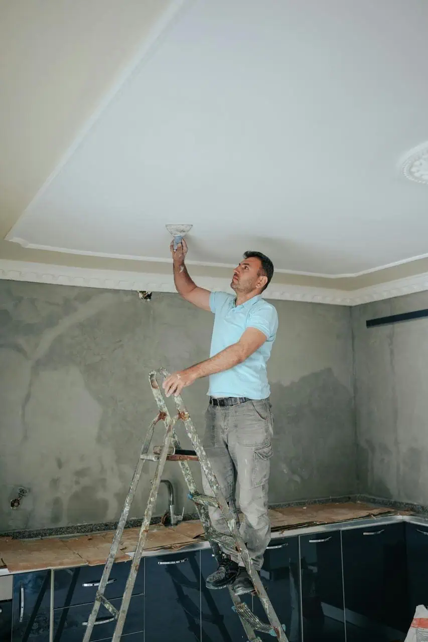 A hotel maintenance technician standing on a ladder, repairing a ceiling light fixture in a modern guest room.