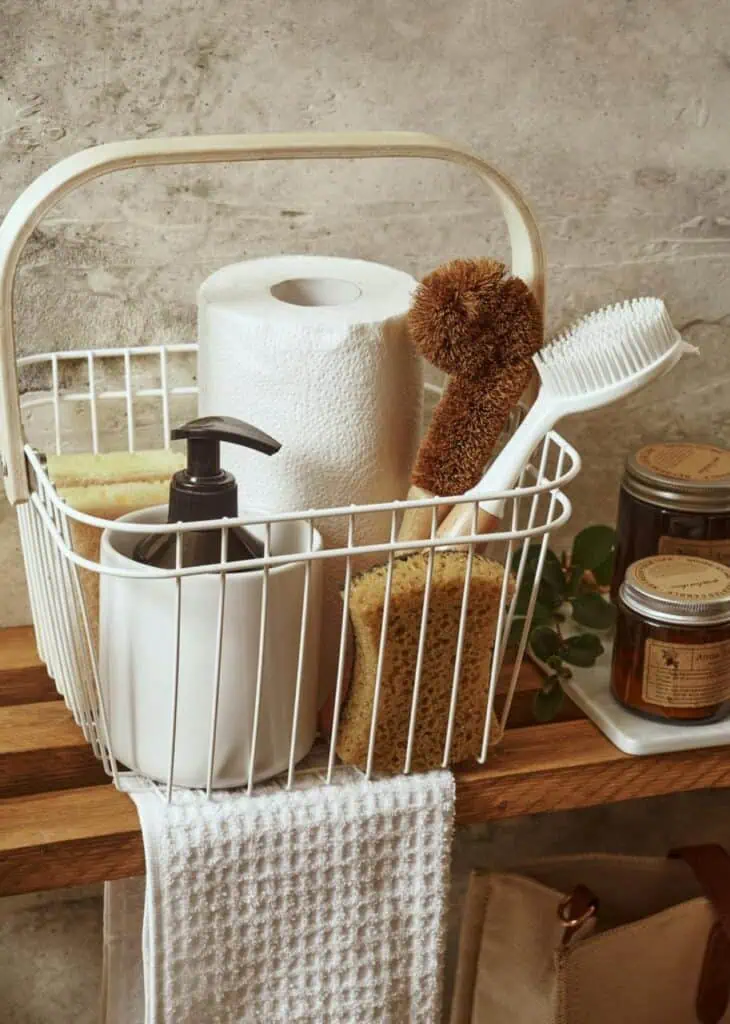 A neatly arranged basket of housekeeping supplies including toilet paper, brushes, soap dispenser, and sponges on a wooden shelf.