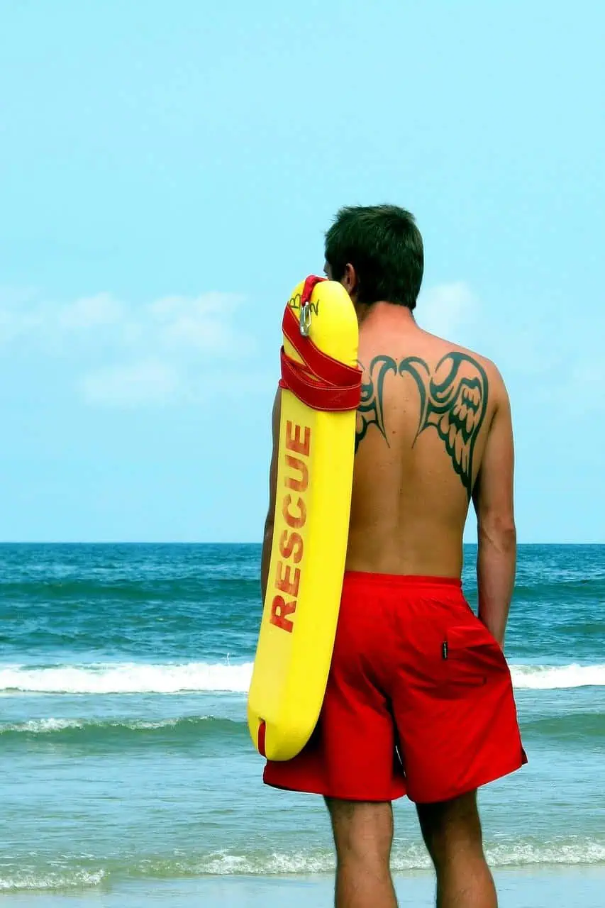 A beach lifeguard in red shorts holding a yellow rescue float, standing watchfully by the ocean to ensure swimmer safety and respond to emergencies.