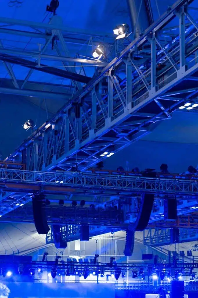 Overhead rig of stage lights and trusses lit in blue—representing the work of lighting technicians in live events and production setups.