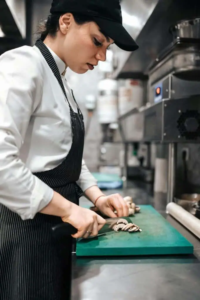 A prep cook slicing fresh mushrooms on a cutting board in a professional kitchen, preparing ingredients for meal service.