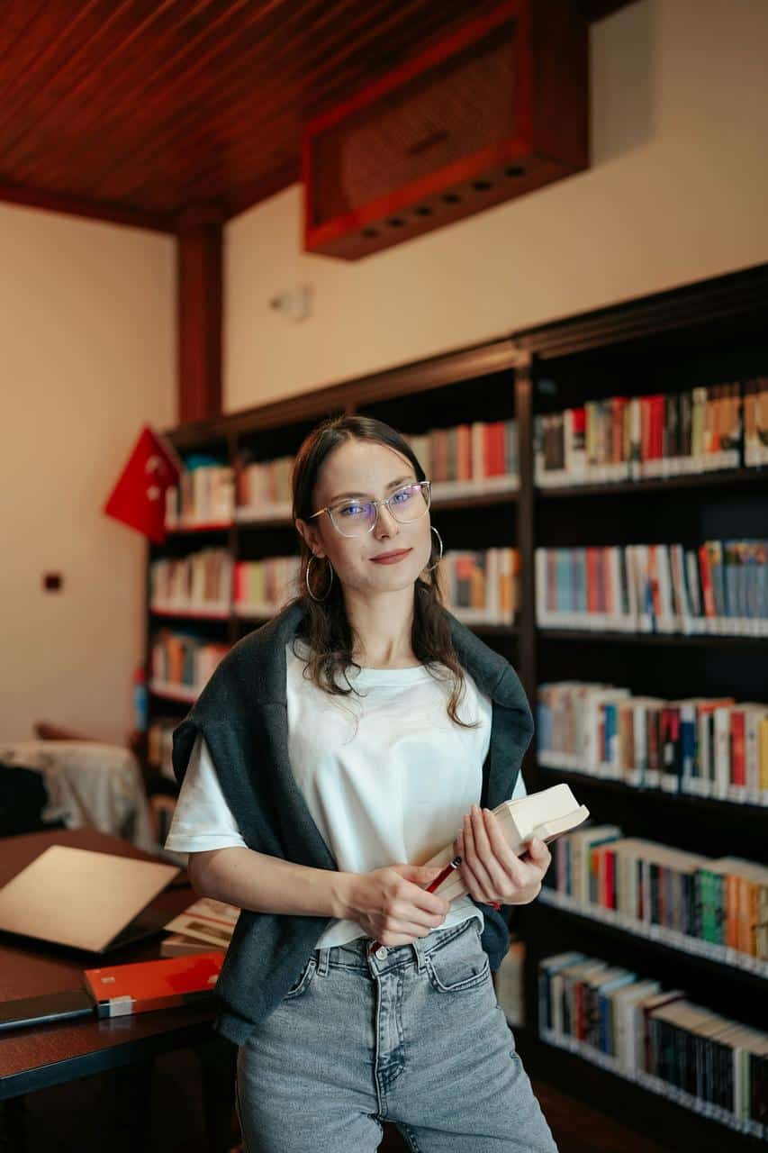 A young public facilities manager standing in a library, holding a book and smiling confidently in front of bookshelves.