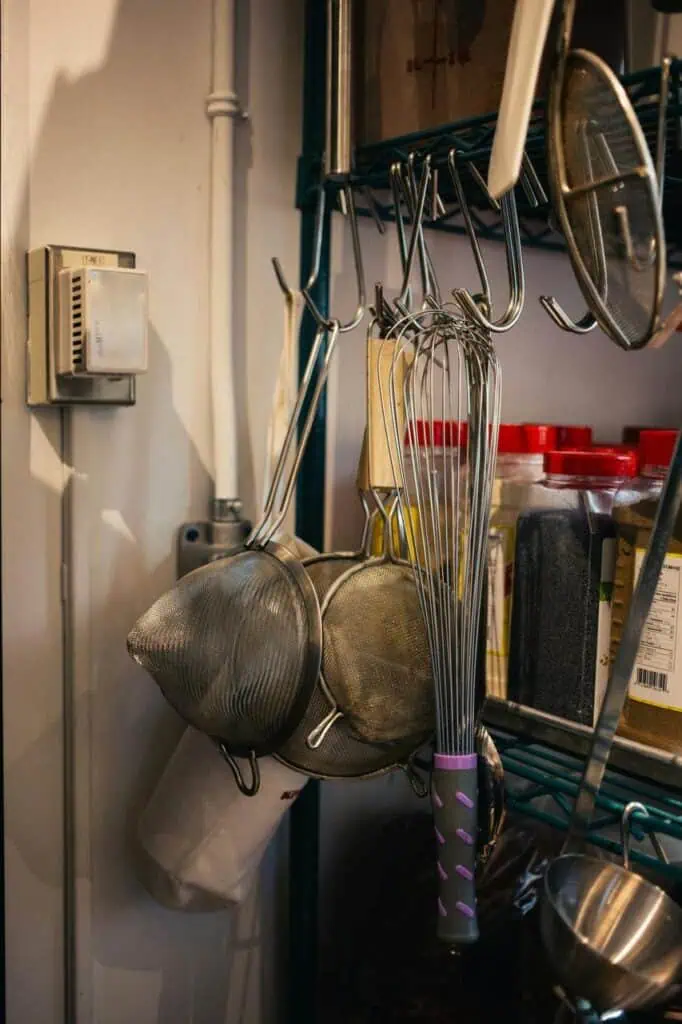 Professional kitchen tools including whisks, strainers, and spices neatly arranged on a restaurant shelf, representing commercial kitchen equipment.