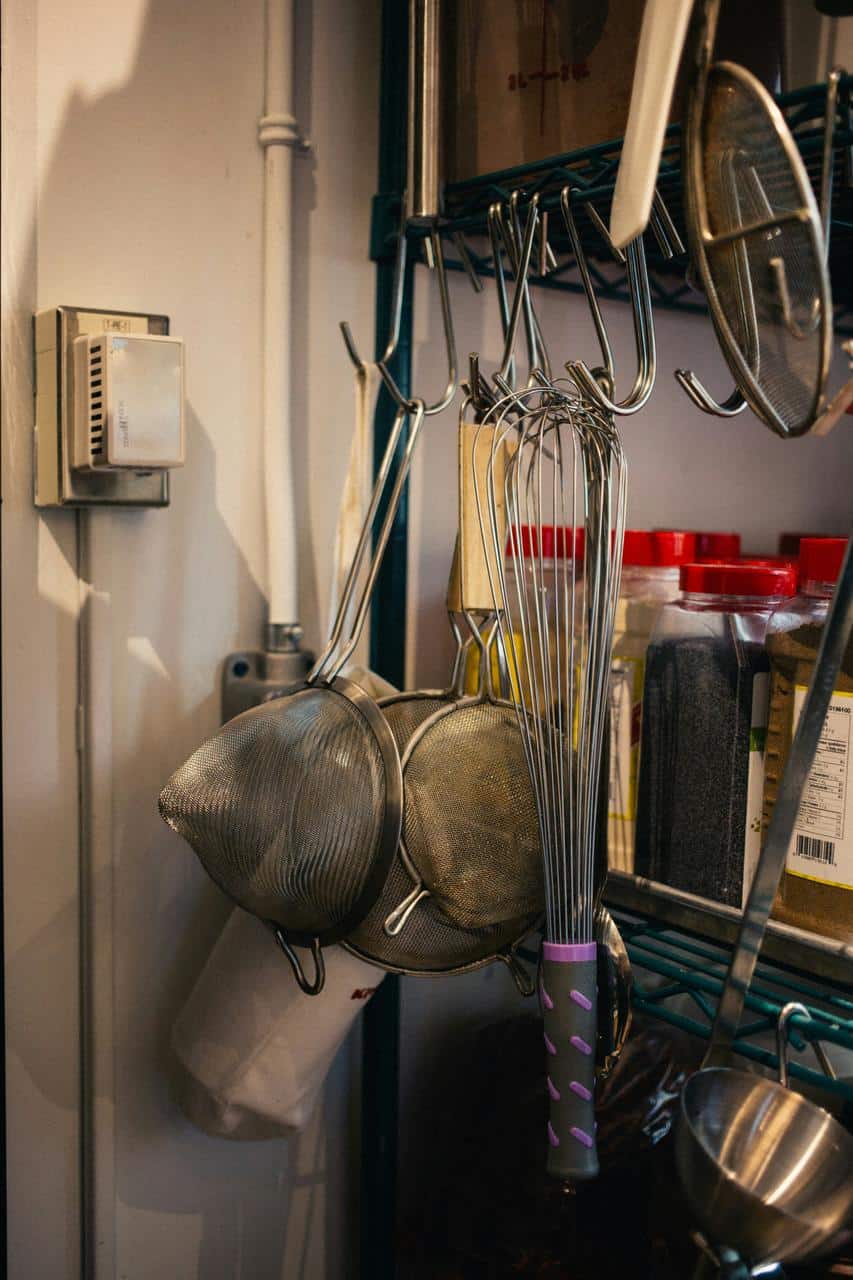 Professional kitchen tools including whisks, strainers, and spices neatly arranged on a restaurant shelf, representing commercial kitchen equipment.