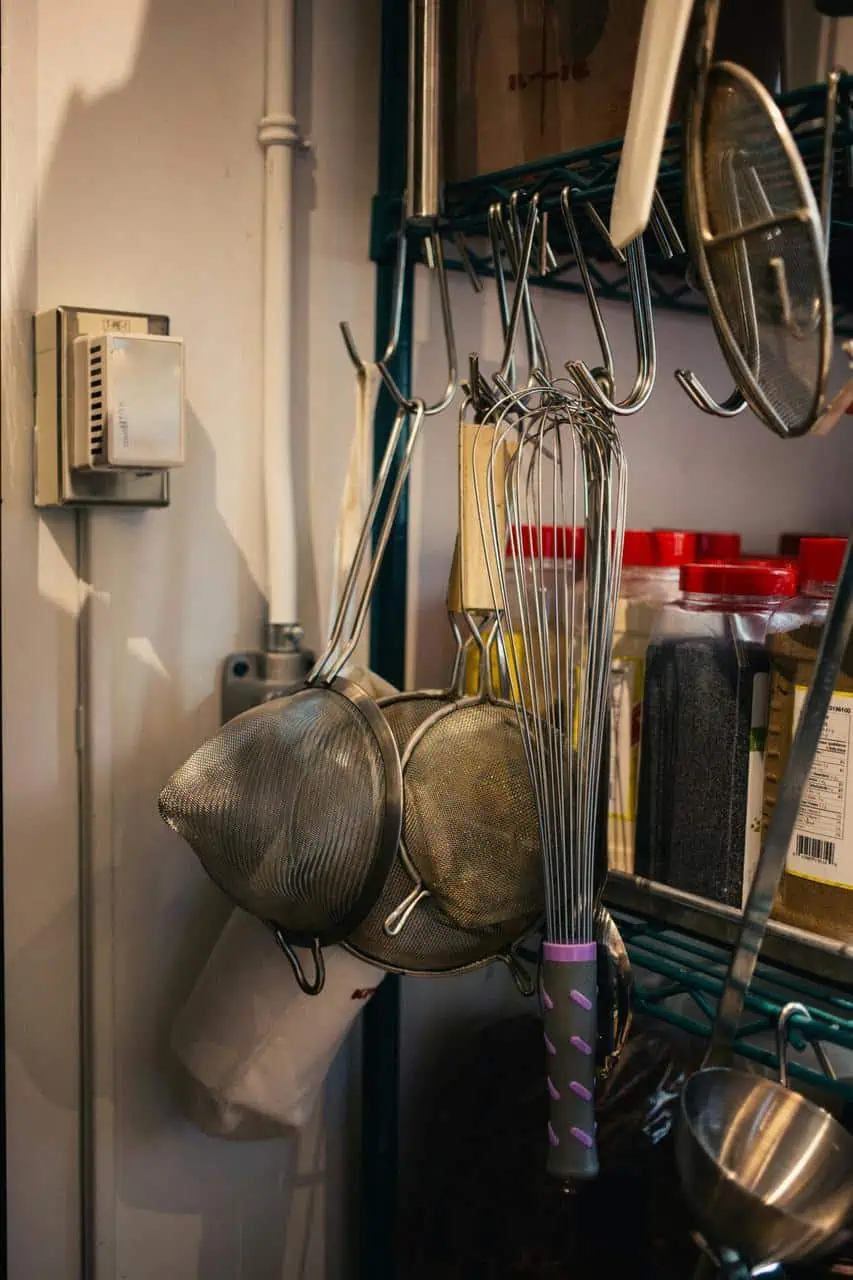 Professional kitchen tools including whisks, strainers, and spices neatly arranged on a restaurant shelf, representing commercial kitchen equipment.