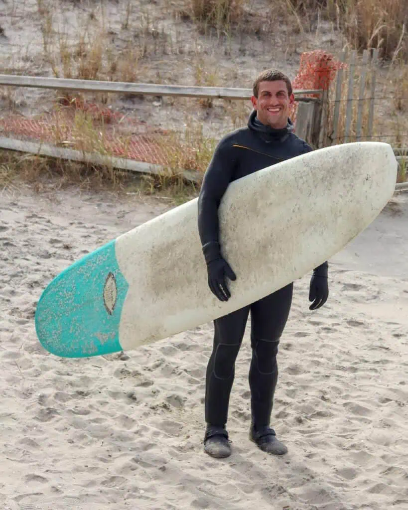 Smiling surfing instructor in a wetsuit holding a surfboard on a sandy beach, ready to coach beginner surfers in ocean safety and wave-riding skills.