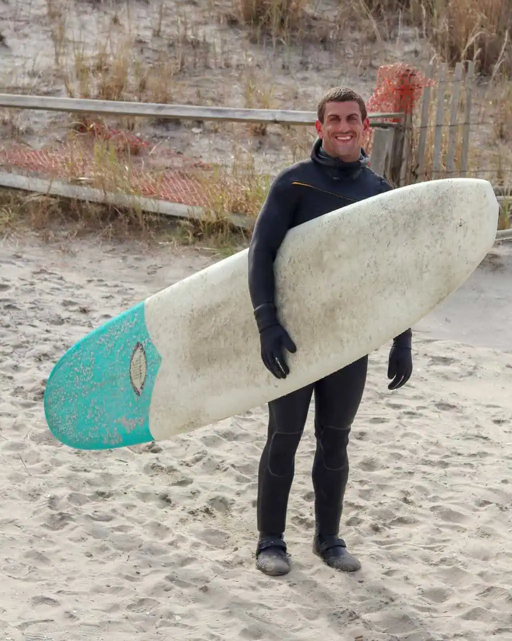 Smiling surfing instructor in a wetsuit holding a surfboard on a sandy beach, ready to coach beginner surfers in ocean safety and wave-riding skills.