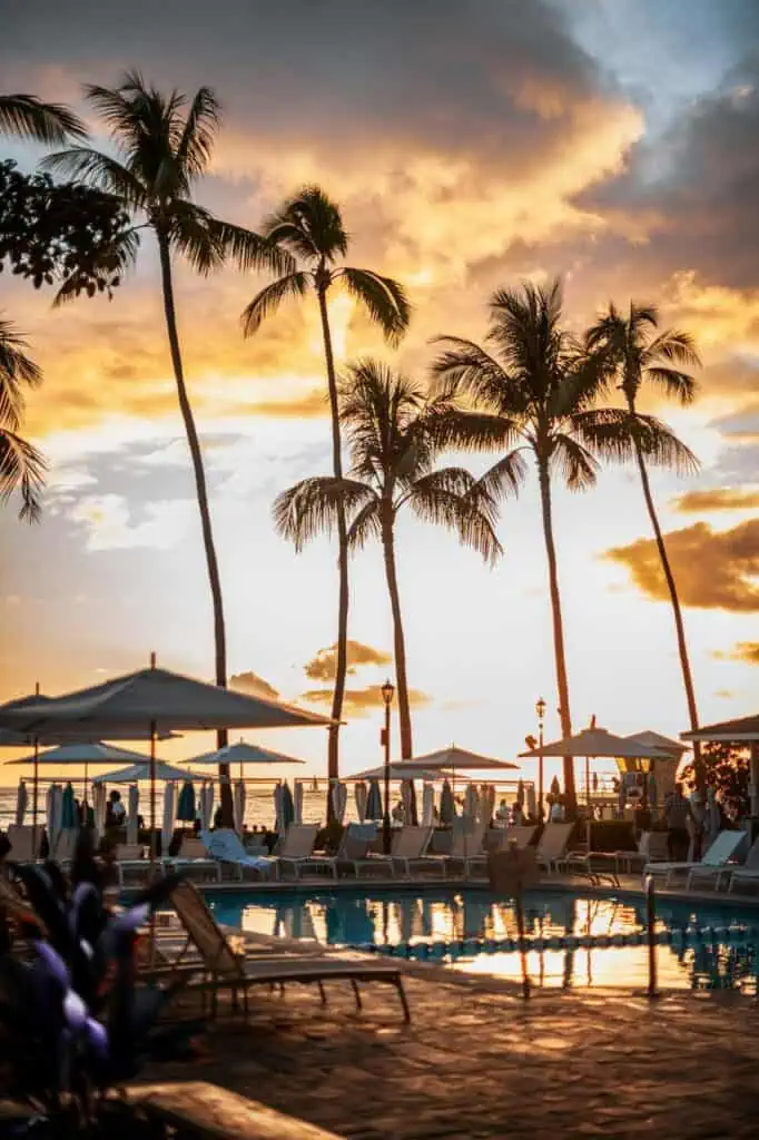 Tropical resort poolside with palm trees at sunset and vacationers relaxing under umbrellas.