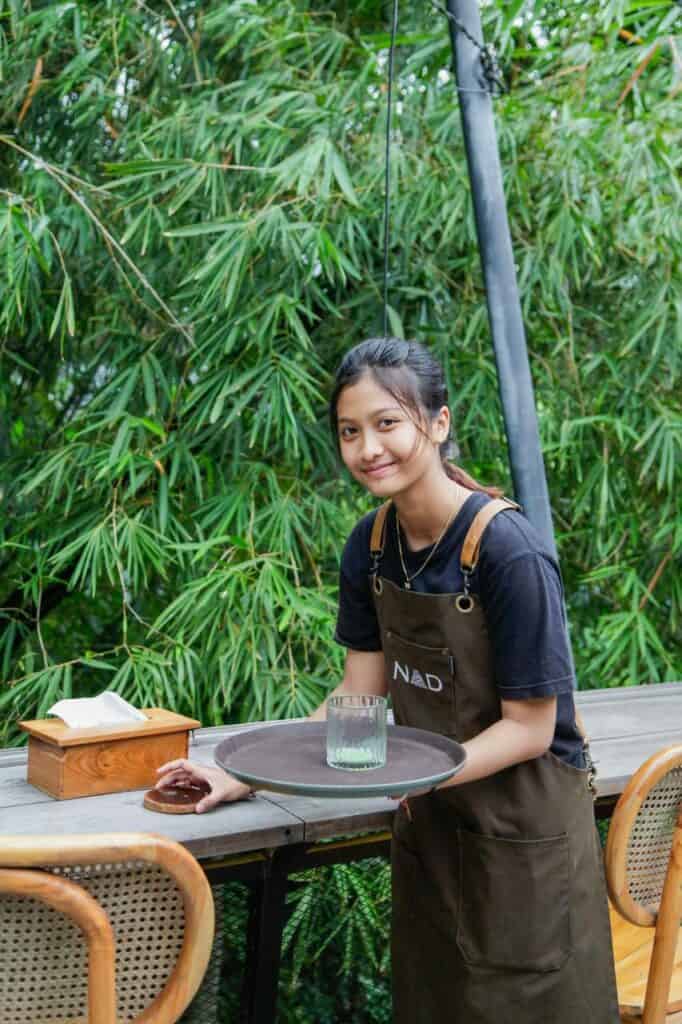 Young waitress serving at an outdoor café, smiling while carrying a tray—representing customer service and entry-level hospitality careers.