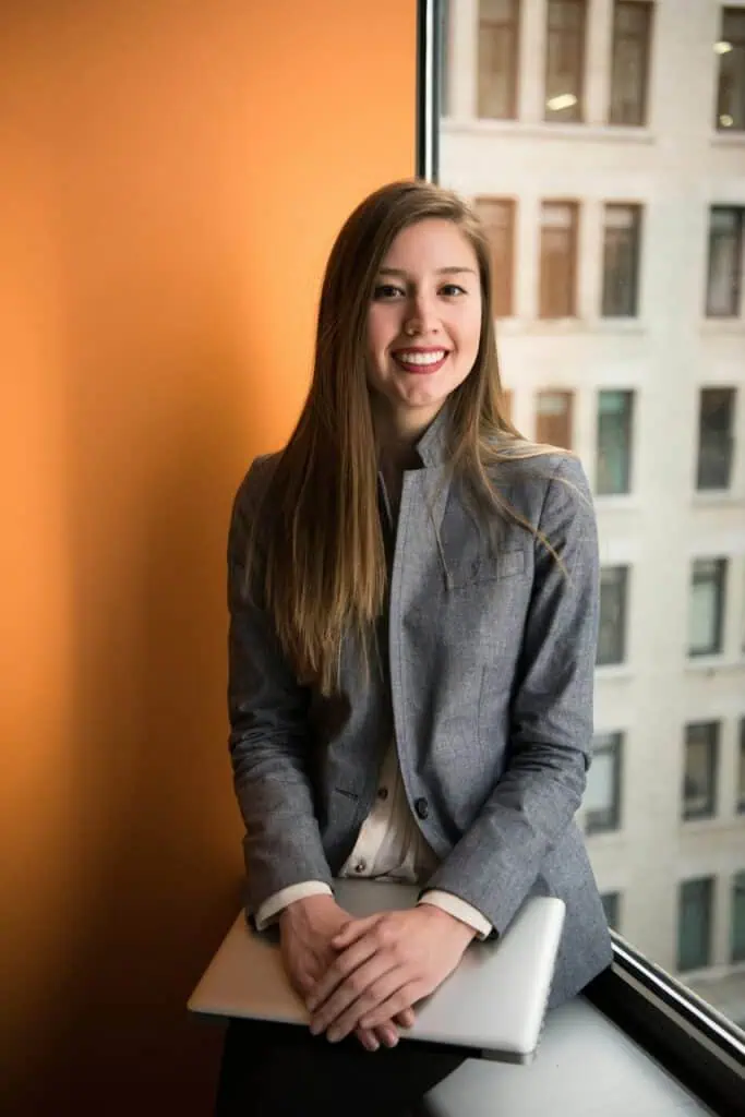 Smiling young professional woman holding laptop seated by office window in business attire