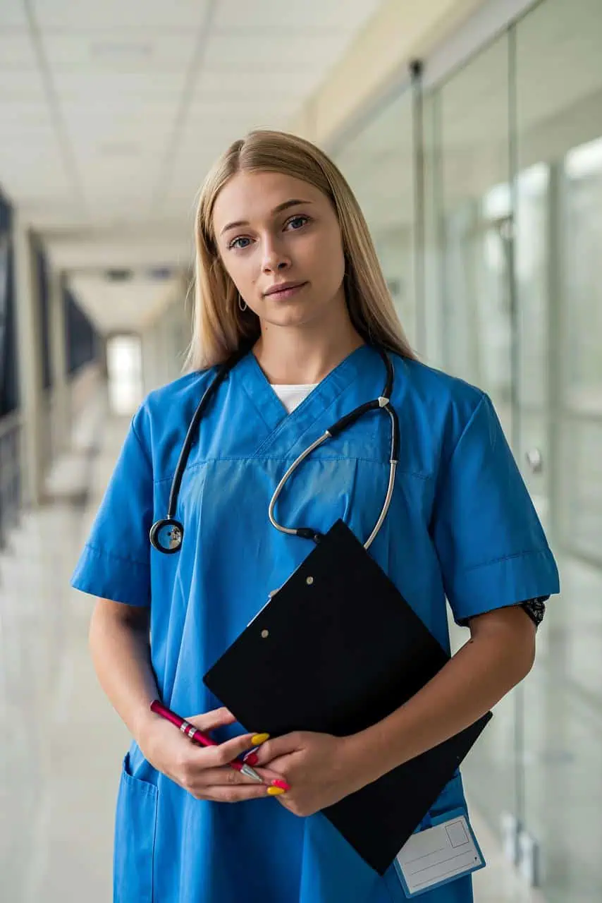Young registered nurse with stethoscope holding clipboard in hospital corridor